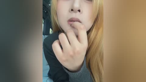 Video of a fair-skinned woman with long blonde hair, wearing a grey sweater, resting her chin on her hand in a contemplative pose. Background shows a car interior.
