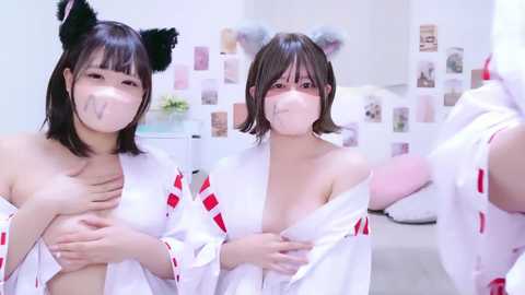 Media: Video of two Asian women in white yukata robes with red trim, wearing cat ear headbands, black cat ears, and face masks, posing in a white room with minimalist decor.