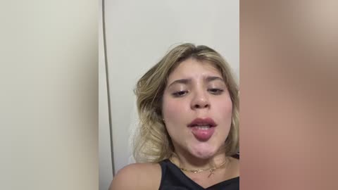 Media: Video of a young woman with fair skin, blonde hair, and full lips, sticking her tongue out playfully against a plain white wall. She wears a black sleeveless top and a gold necklace.
