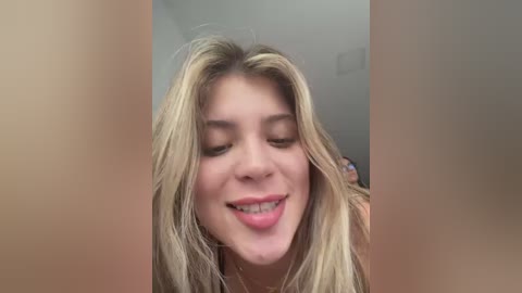 Video of a young woman with fair skin and long blonde hair, smiling playfully with her tongue out. The blurred background suggests an indoor setting.