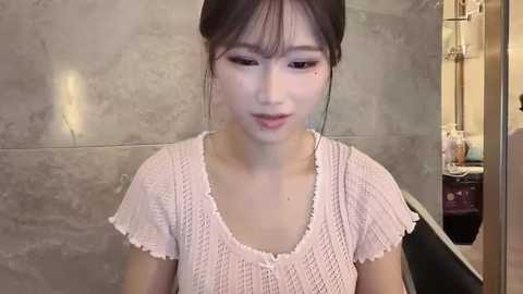 Video of an East Asian woman with fair skin, straight black hair tied back, wearing a light pink knitted top, standing in a modern bathroom with gray tiled walls and a sink visible in the background.