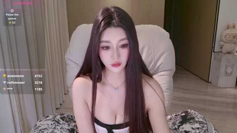 Video of an East Asian woman with long, straight black hair and fair skin, wearing a strapless black and white lingerie top, sitting on a bed with a white blanket.