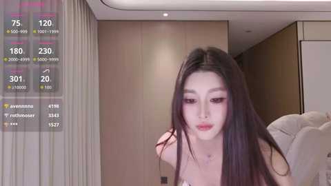 Media: Video of an East Asian woman with long black hair, wearing a sleeveless top, sitting in a modern room with beige curtains and wooden walls, displaying social media metrics overlay.