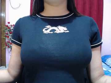 Video of a woman with medium skin tone wearing a tight, dark navy T-shirt with white trim and a white dragon logo. She has long, straight black hair and is standing indoors, with a red curtain and decorative plant in the background.