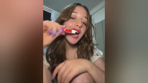 Media: Video of a young woman with light skin and long brown hair, wearing a floral shirt, brushing her teeth in a dimly lit bathroom with beige walls.