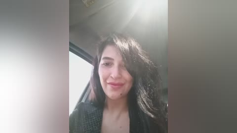 A video of a young woman with long, dark hair, fair skin, and a slight smile, wearing a dark sweater, taken from inside a car with sunlight streaming in.