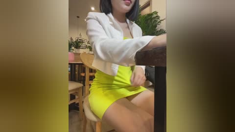 Media: A video of an East Asian woman with shoulder-length black hair, wearing a white cardigan over a bright yellow mini dress, sitting at a wooden table in a cozy indoor setting.
