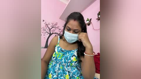 Video of a young woman with medium skin tone and black hair, wearing a blue floral dress and a blue surgical mask, sitting in a pink room with tree decals and a red sofa.