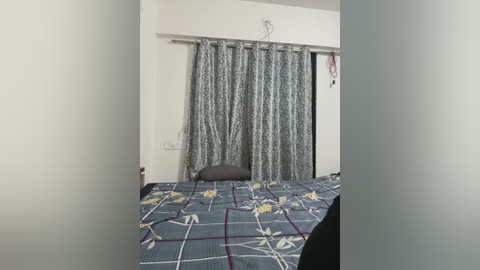 Video of a simple bedroom with a blue and white floral-patterned bedspread, gray curtains, and a small black chair. The walls are white, and the room is well-lit.
