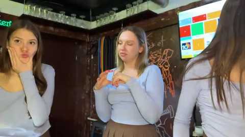 Video of three young women, two with long hair, in casual light blue tops, making silly faces in a dimly lit bar with a TV screen in the background.