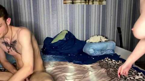A video captures a nude woman with large breasts and a tattoo on her chest, standing beside a nude man with a tattoo on his chest. They're in a bedroom with striped wallpaper and disheveled blue bedding.