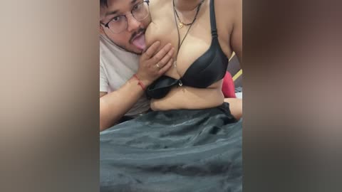 Video of a man with glasses and mustache licking the nipple of a woman in a black bra, lying on a bed with a grey blanket.