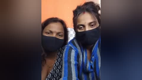 Video of two South Asian women wearing black face masks and blue-patterned traditional clothing. Background features an orange wall and a dark gradient.
