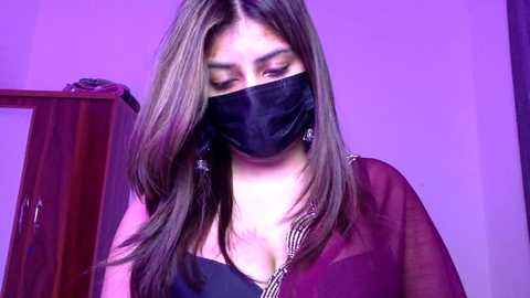 Video of a woman with long brown hair, wearing a black face mask, black bra, and sheer maroon shawl, standing against a purple-lit background.