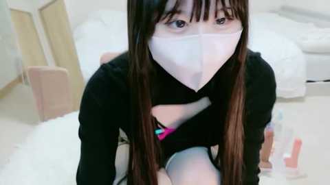 Video of an East Asian woman with long brown hair, wearing a black top and white mask, kneeling on a white carpet, in a minimalist bedroom with white furniture and bottles.