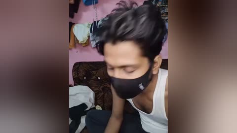 Video of a man with dark hair, wearing a black face mask and white tank top, sitting on a brown couch in a cluttered room with pink walls.