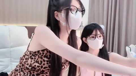 Video of two Asian women with long black hair, wearing white face masks, leopard-print dresses, and glasses, sitting on a white leather couch, in a modern, beige-walled room.