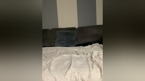 A video of a beige and gray striped wall with a plush gray sofa, a fluffy white blanket, and a blue throw pillow.