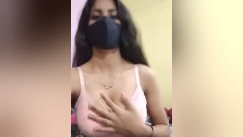 Video of a woman with long black hair, wearing a black face mask, white spaghetti strap top, and gold necklace, covering her chest with her hands.