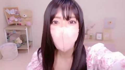Video of an Asian woman with long black hair, wearing a white face mask, pink floral shirt, and a light-colored room with a soft pink carpet, shelves, and stuffed toys.