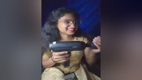 Video of a smiling young woman with curly black hair, wearing glasses and a brown patterned dress, holding a black power drill against a dark blue backdrop.
