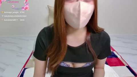 Media: Video of a woman with long auburn hair wearing a black top and pink face mask, seated on a colorful quilted blanket in a minimalistic, white-walled room.
