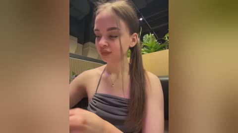 Video of a young woman with fair skin and long brown hair tied in a ponytail, wearing a gray tank top, sitting indoors near a plant. She has a contemplative expression.