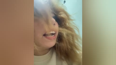 Media: A close-up video of a young woman with curly, shoulder-length blonde hair, smiling widely, revealing her teeth. The background is a plain, light-colored wall.