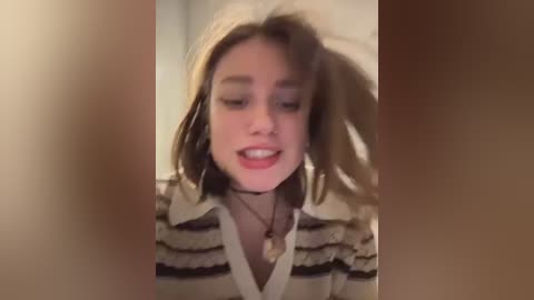 Video of a young woman with light skin and light brown hair, wearing a striped shirt and a choker necklace, laughing with her mouth open, blurred background.