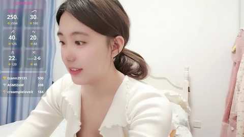 Video of a young East Asian woman with straight brown hair, wearing a white top, in a bedroom with a bed, pink towel, and a blue-striped curtain in the background.