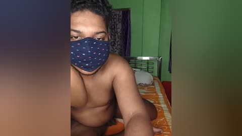 Video of a topless Black man with short curly hair, wearing a blue mask with white dots, sitting on a bed with orange and white bedding in a dimly lit room with green walls.