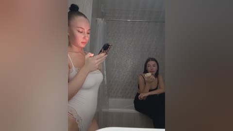Video of a pregnant woman in a white lace bodysuit texting while another woman in a black dress eats a banana in a bathroom with beige tiled walls.