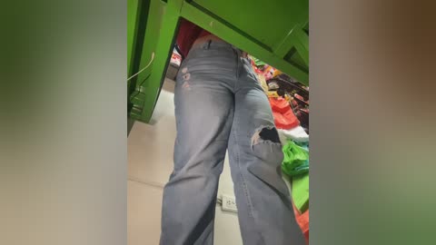 A video captures a person's lower torso from a low angle, wearing distressed blue jeans with a ripped pocket and a green shirt. The background features a cluttered closet with various colorful clothes.