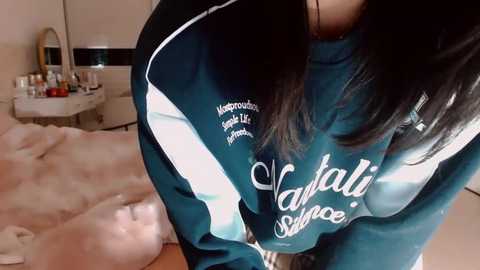 Video of a woman with long black hair wearing a teal and black athletic jacket with \"Valley Service\" text, standing in a dimly lit bedroom with a white dresser, mirror, and bed in the background.
