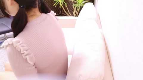 A video shows an Asian woman with long black hair, wearing a pink, ruffled top, leaning against a white wall with green plants in the background.