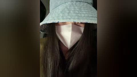 A video of a person with long, straight brown hair, partially obscured by a light blue bucket hat. They wear a pink surgical mask, covering their nose and mouth, and a light blue shirt with a subtle pattern. The background is blurred, focusing attention on the face.