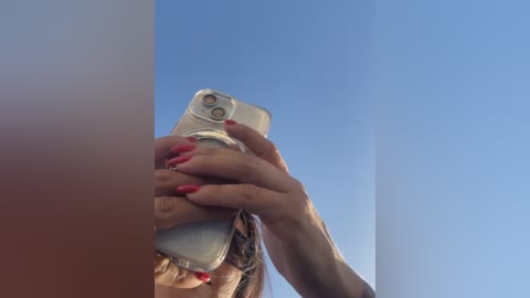 Video of a person holding a silver iPhone with red nails, capturing a selfie against a clear blue sky.