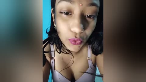 Media: Video of a young South Asian woman with medium brown skin, wearing a lavender bra, blowing a kiss against a blue background.
