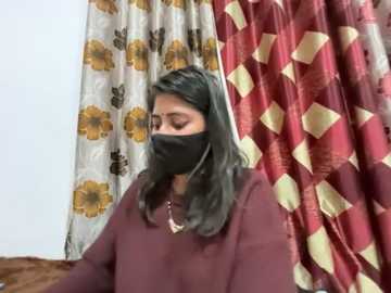 Video of a South Asian woman with medium skin tone, wearing a black mask and maroon top, standing in a room with floral-patterned curtains.