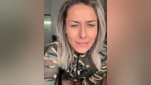 Video of a woman with long, gray hair, wearing a camo-patterned, zip-up jacket, in a modern kitchen.