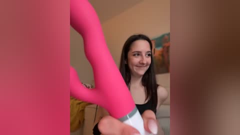 Video of a smiling woman with long dark hair, wearing a black tank top, holding a pink sex toy in a beige room with a bed and abstract wall art.