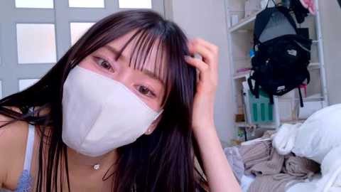 Media: Video of an East Asian woman with long black hair, wearing a white mask, lying on a bed with disheveled sheets, surrounded by cluttered shelves and a black backpack.