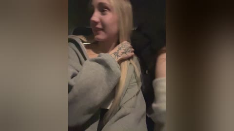 Video of a young blonde woman with a tattooed arm, wearing a grey hoodie, looking to the side while talking on a black phone, dimly lit background.