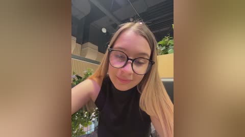 Video of a young, fair-skinned woman with long blonde hair and glasses, wearing a black top, standing in a modern, well-lit room with green plants in the background.