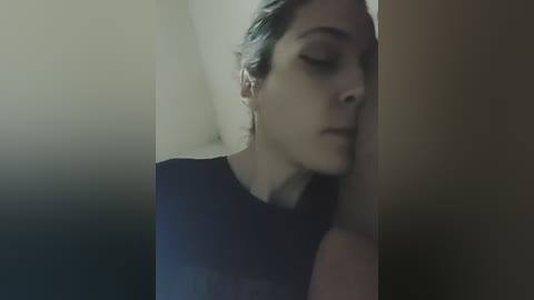 Video of a young woman with pale skin, blue eyes, and light brown hair, leaning against a beige wall. She wears a dark, short-sleeved top, eyes closed, appearing tired.