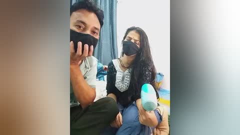 A video of a man and woman wearing face masks, smiling, in a room with blue curtains. The woman holds a blue ice pack, and the man has a black mask and is holding a bottle.