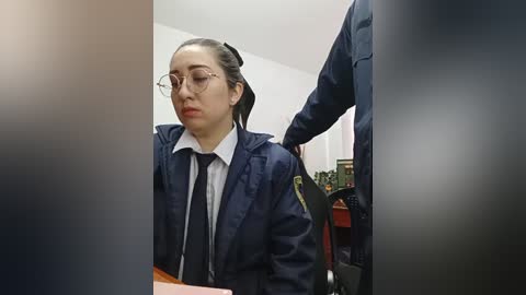 Video of a young Asian woman with glasses, wearing a navy jacket over a white shirt and black tie, seated in an office chair, surrounded by blurred figures.