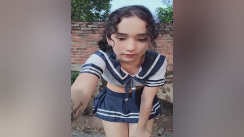 Video of a young person with curly dark hair, wearing a sailor schoolgirl outfit with a white and navy blue skirt, crouching outdoors by a brick wall.