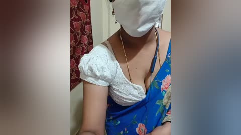Video of a woman with medium skin tone, wearing a blue floral dress with a white lace top, a white face mask, and a gold necklace. Background shows a red floral curtain.