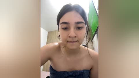 A video of a young Latina woman with medium skin tone, straight dark hair, and a strapless blue top, seated in a brightly lit, minimalist room with white walls and a green wall-mounted television.